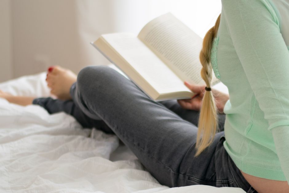A young woman reads a book by bedside lamp in a cozy bedroom setting at night.