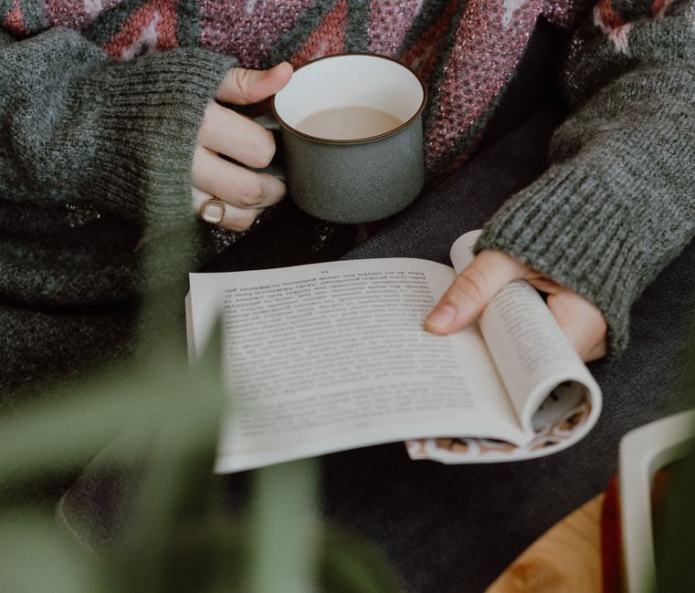 Person holding a coffee mug while reading a book indoors, exuding a warm and cozy atmosphere.